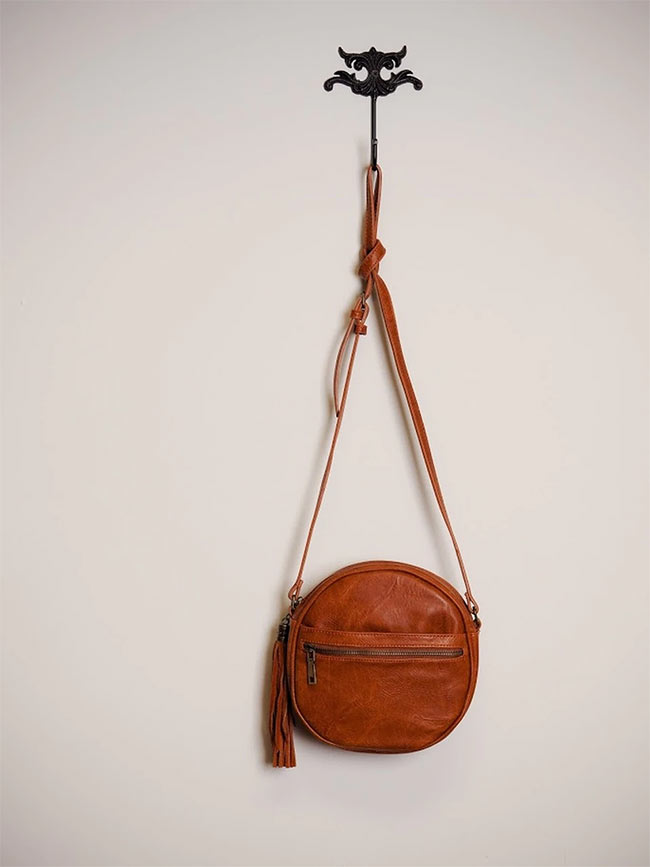 A brown, circular crossbody bag hangs on a wall hook