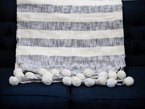  A white and gray striped pom-pom throw blanket modeled on a couch as an example of rustic spring decor.