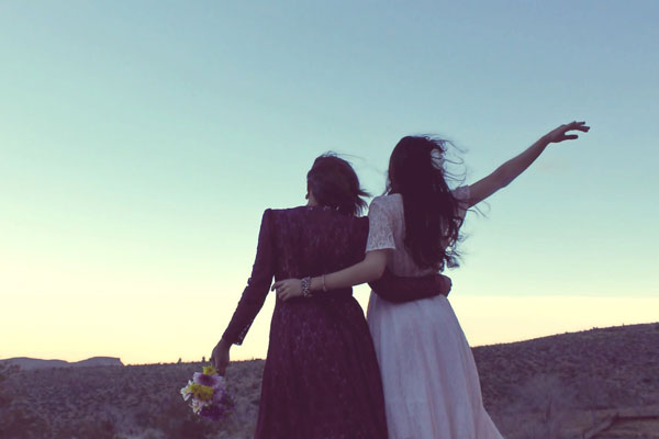 Two women wearing modern hippie style-clothes hold on to each other in the desert.