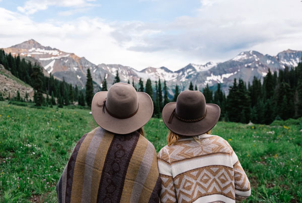 Two women sporting modern hippie style sit together in a field overlooking the mountains.